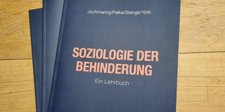 Drei Bücher liegen auf einem Tisch. Titel des oberen Buches ist "Soziologie der Behinderung: Ein Lehrbuch" in orangener Schrift auf schwarzem Grund. Darüber die NAmen der Autor*innen Jochmaring, Pelka, Stange und York.