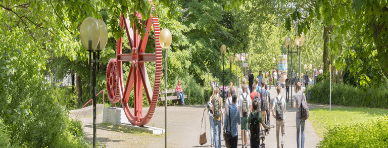 Kopfbild Studenten rote Zahnräder Campus Nord Studenten im Sommer auf dem Campus Nord, rote Zahnräder im Hintergrund