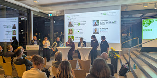 Auf dem Podium stehen fünf Diskutierende sowie zwei Moderierende an einem Rednerpult der TU Dortmund. Auf der Leinwand werden ihre Namen und Institutionen angezeigt.