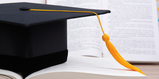 Ein schwarzer Doktorhut mit einer orangenen Quaste liegt auf einem aufgeschlagenen Buch.