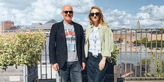 Ein Mann und eine Frau mit Konferenz-Lanyards stehen lächelnd auf einer Dachterrasse, im Hintergrund die Skyline von Helsinki mit Hafen und blauem Himmel mit Wolken.