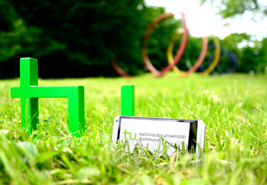 TU-App Ein Smartphone, das neben dem TU-Logo im Gras steht, im Hintergrund sind die Spektralringe zu sehen.