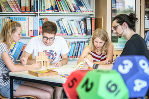 Mathedidaktik-Werkstatt Vier Personen sitzen an einem Tisch und beschäftigen sich mit Bausteinen, die auf dem Tisch liegen. Im Vordergrund sieht man bunte vieleckige Würfel. Im Hintergund steht ein Regal voller Bücher und Ordner.